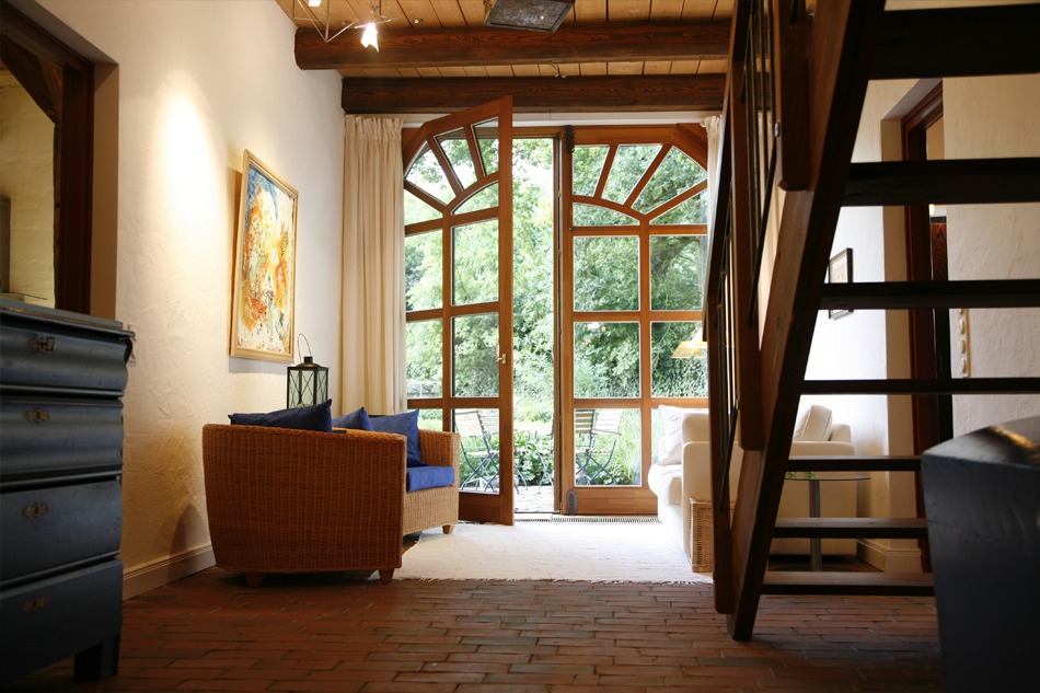 FeWo Harms Hoff – Wohnzimmer Blick in das Wohnzimmer mit Steinfußboden und Teppich, Holzbalken an der Decke und offener Holztreppe, offene Terrassentür mit Fenstern bis zum Boden in der Reetdach-Fewo Harms-Hoff in Hanstedt-Schierhorn